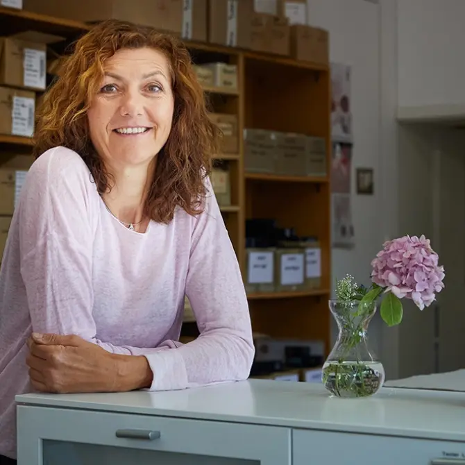 Frau lehnt auf einem Tisch mit einer Blumenvase und einer Kosmetiktube darauf. 