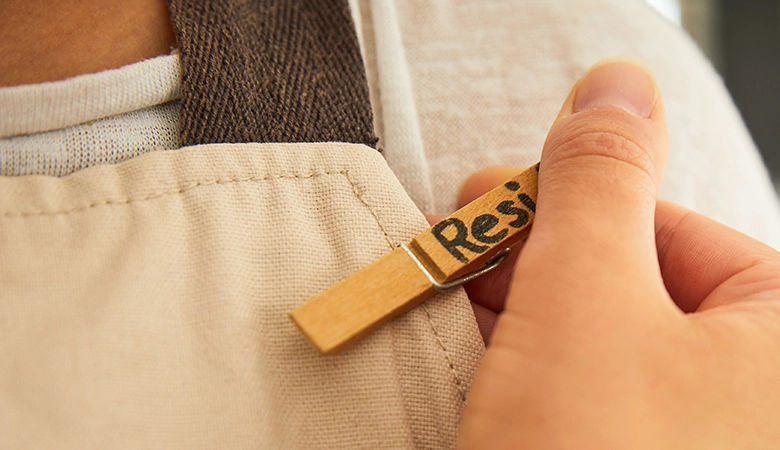 Eine Hand heftet eine Büroklammer mit der Aufschrift "Resi" an eine Schürze. 