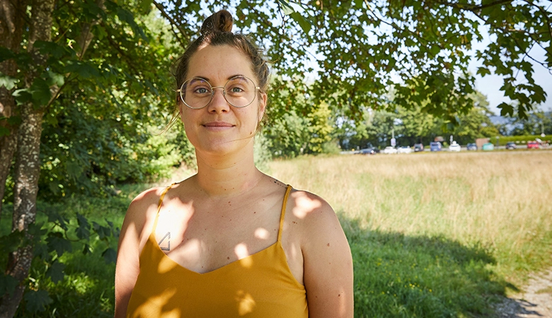 Eine junge Frau steht an einem Feldweg neben einem Baum und lächelt. 