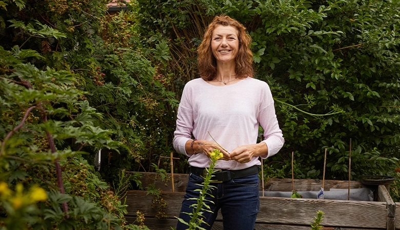 Frau steht in einem Garten mit einem Halm in der Hand und lächelt. 