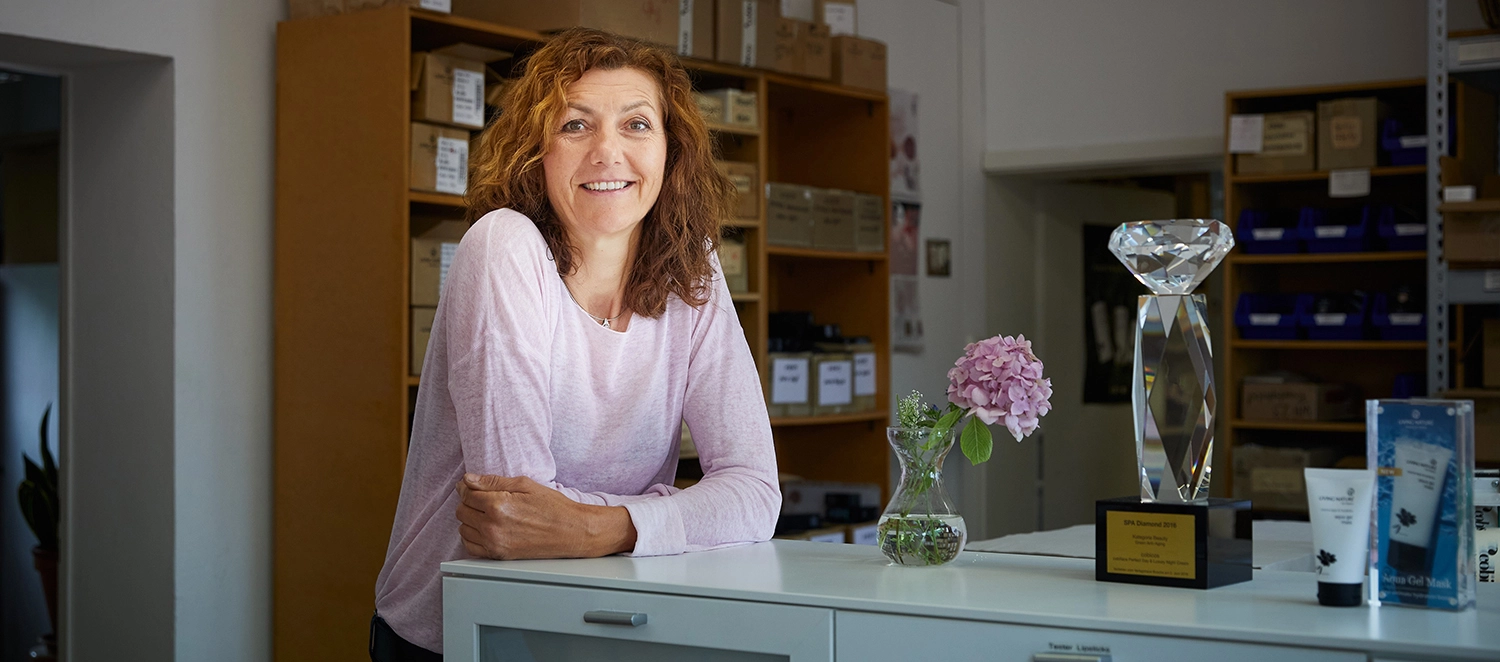 Frau lehnt auf einem Tisch mit einer Blumenvase und einer Kosmetiktube darauf. 