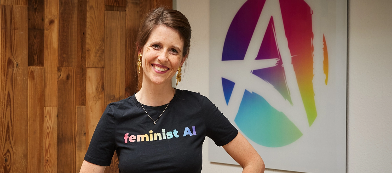 Eine junge Frau lächelt in die Kamera. Sie trägt ein T-Shirt, auf dem "feminist AI" steht. 